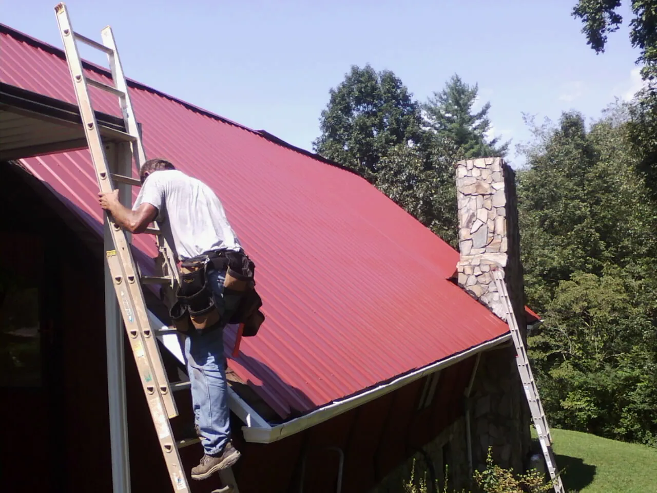 Roofer on a ladder beside a red metal roof and stone chimney — Soffit & Fascia Repair by Roofing Brothers in Dinwiddie